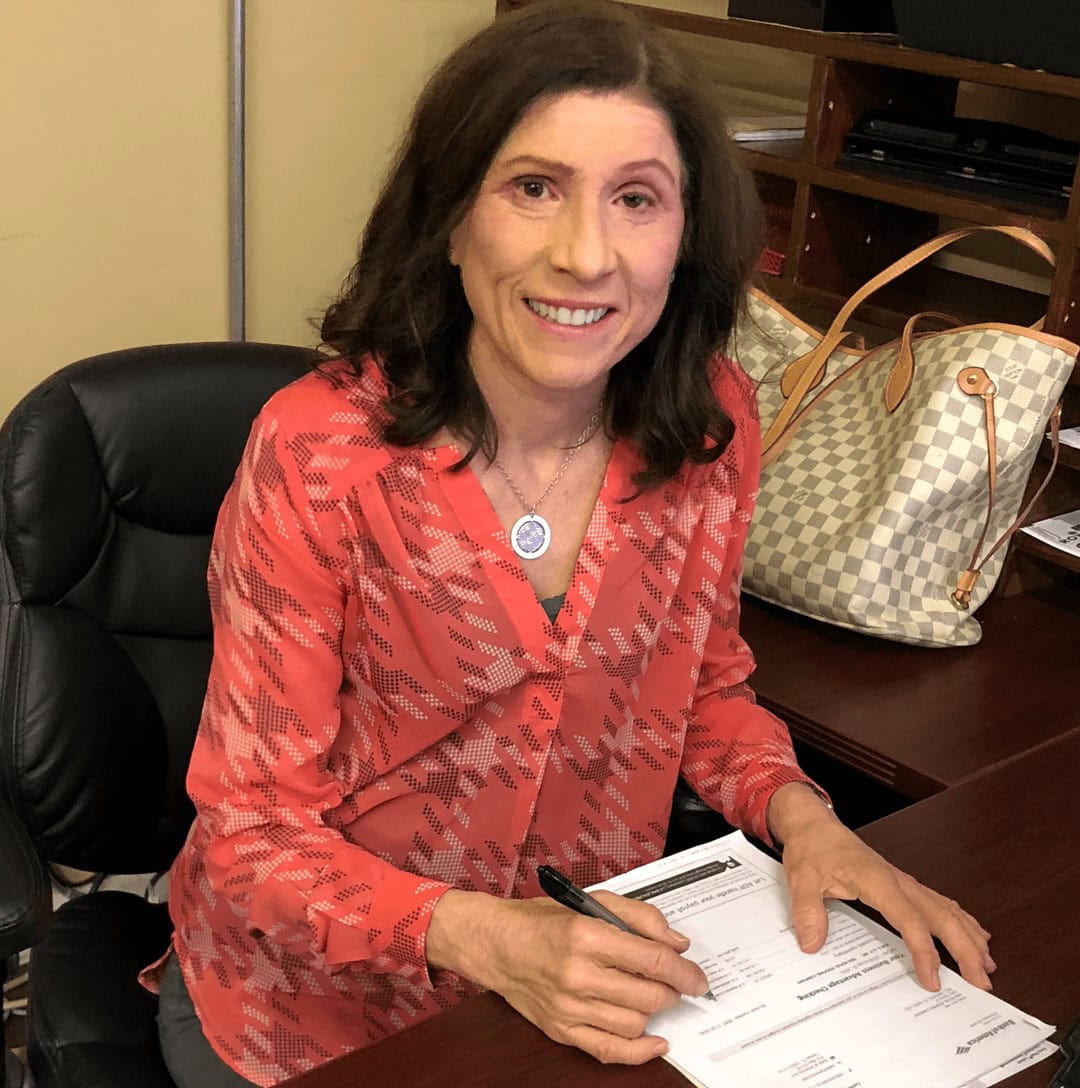 A woman sitting at a desk, smiling at the camera while filling out paperwork with a pen. A handbag is on the adjacent desk.