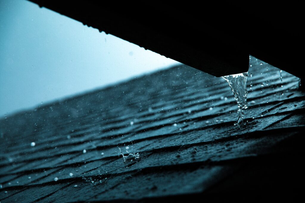 Rainwater pours off the edge of a roof onto dark shingles, droplets splashing in a moody blue light—capturing the expertise of a roofing contractor in Orange & Los Angeles County, CA on a classic rainy day.