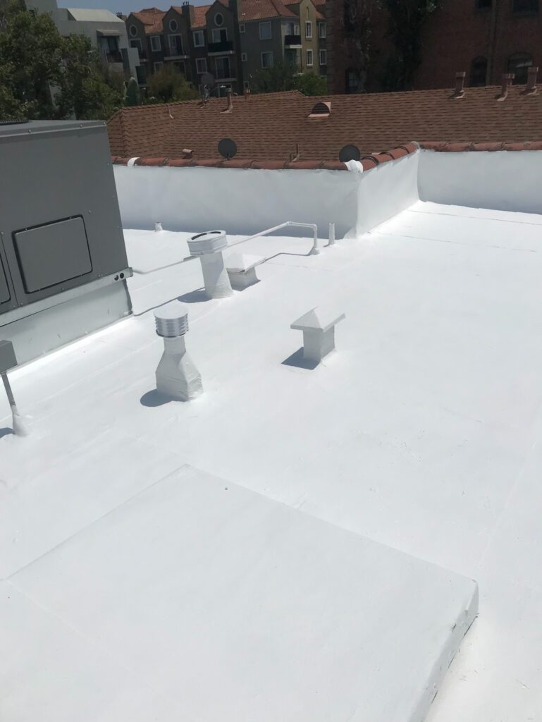 A flat white-painted rooftop with several ventilation pipes and units, bordered by a low white wall. A roofing contractor in Orange & Los Angeles County, CA could maintain this space, surrounded by nearby red-tiled roofs under a clear sky.