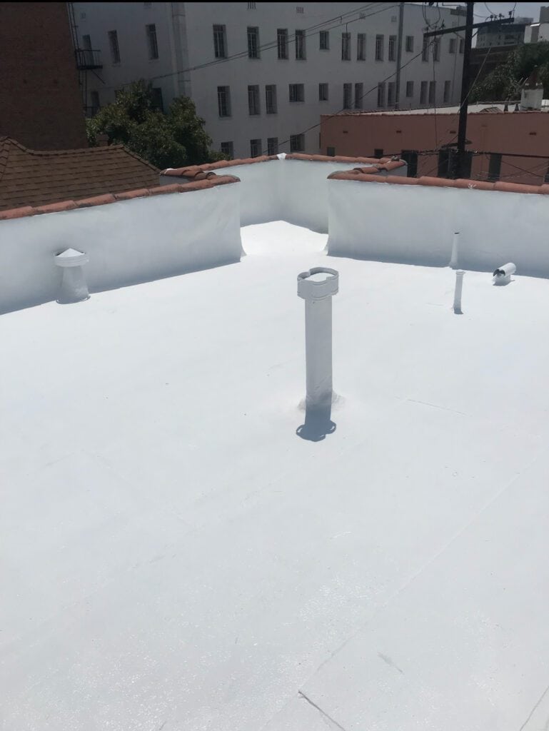 A flat white rooftop with several vent pipes and surrounding short walls, located in an urban area with buildings and rooftops visible in the background under bright sunlight, maintained by a trusted roofing contractor in Orange & Los Angeles County, CA.