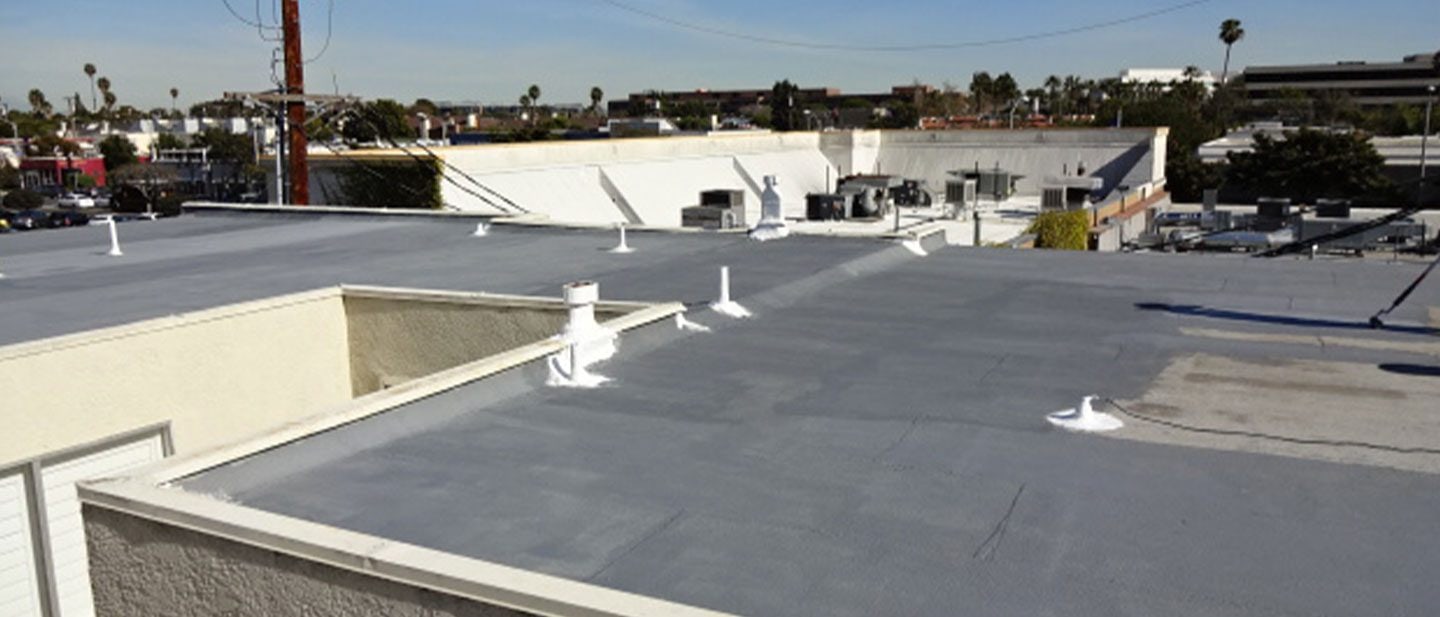 Flat commercial building rooftop with gray coating and several white vent pipes, maintained by a roofing contractor in Orange & Los Angeles County, CA; surrounded by other buildings, palm trees, and a clear blue sky in the background.