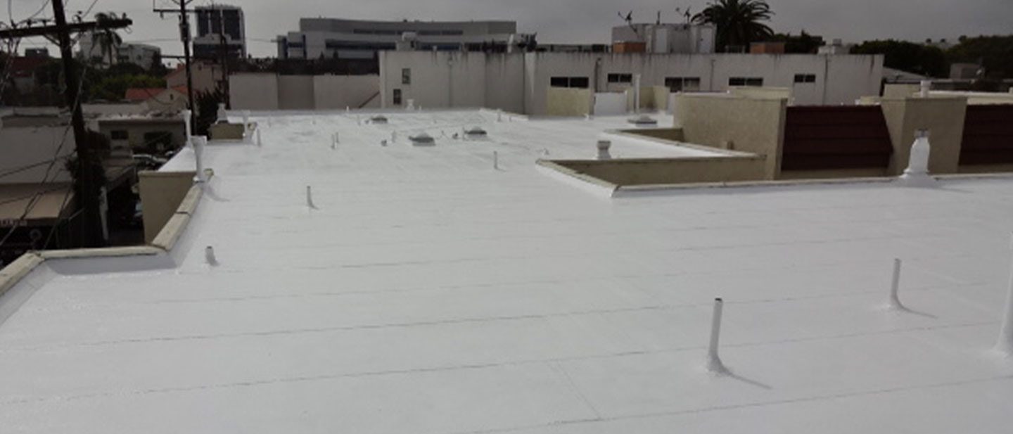 A flat white rooftop with multiple pipes and vents, serviced by a trusted roofing contractor Orange & Los Angeles County, surrounded by other buildings and some trees in the background under an overcast CA sky.