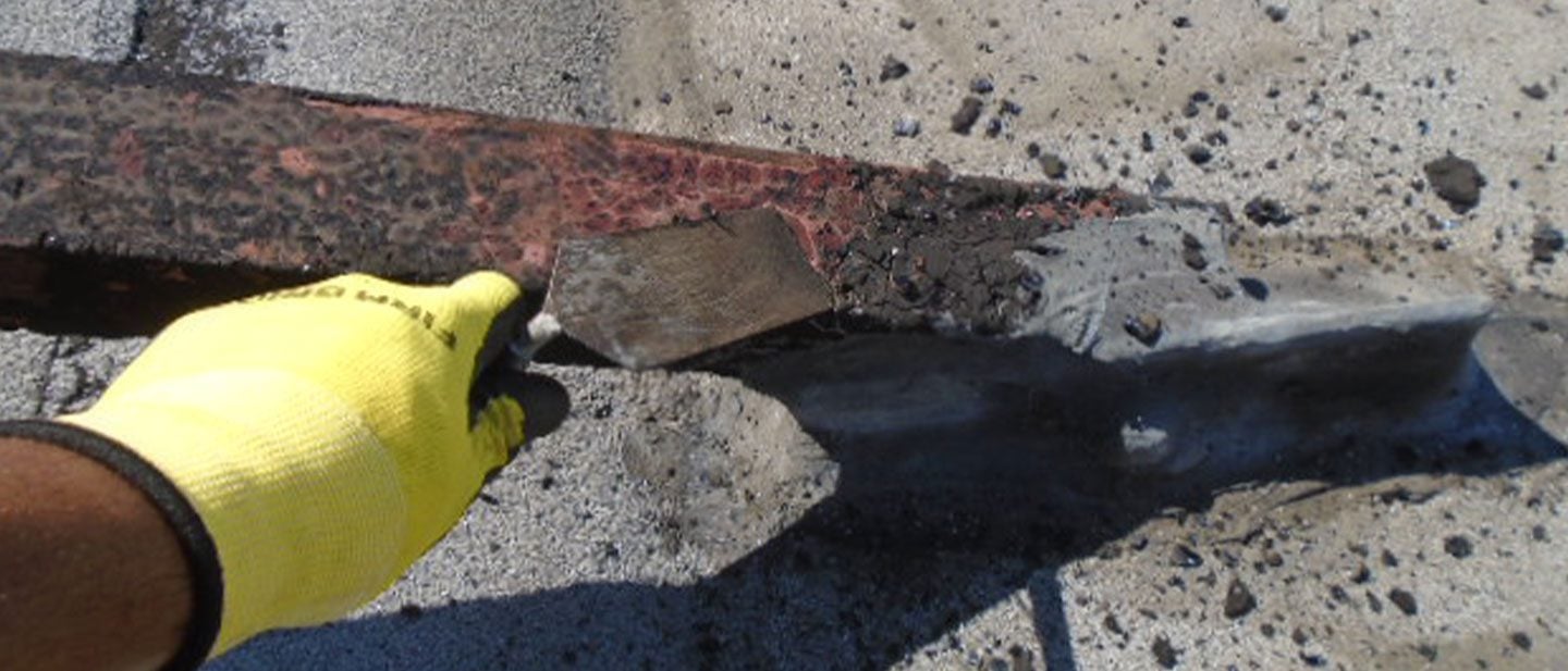 A roofing contractor in Orange & Los Angeles County, CA, wearing a yellow glove, uses a metal scraper to remove old roofing material from the edge of a roof, revealing debris and the exposed surface underneath.