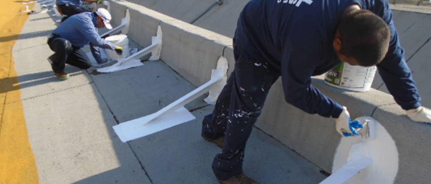 Workers in blue shirts and white gloves, possibly from a CA roofing contractor Orange & Los Angeles County, are painting metal rail supports white along a road or bridge barrier. Paint cans and brushes are visible, with paint splatters on the concrete.
