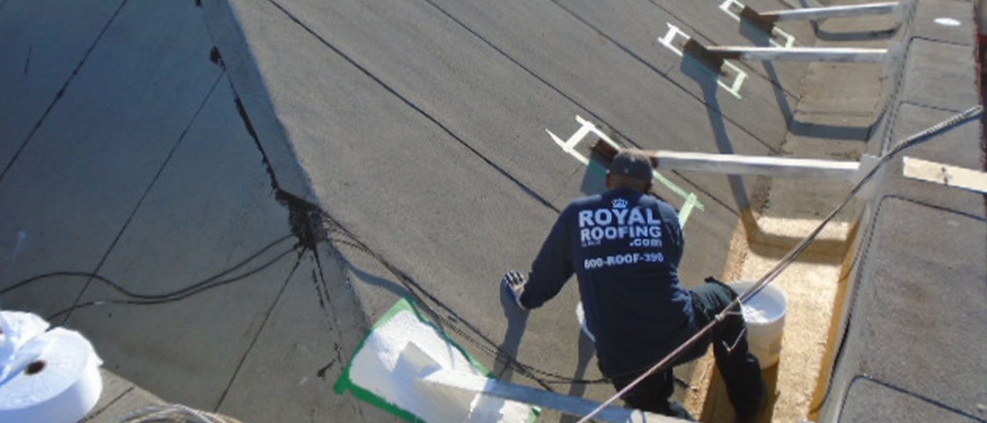 A worker in a "Royal Roofing" shirt, representing a top roofing contractor in Orange & Los Angeles County, CA, applies roofing materials on a sloped roof using tools and buckets, with white vent pipes and wooden supports visible.