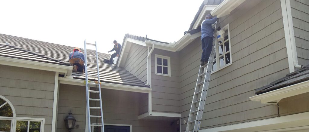 Three workers are painting and repairing a light gray house in CA; two, possibly roofing contractor Orange & Los Angeles County specialists, are on the roof by ladders while one paints near an upper window. The sky is bright and clear.