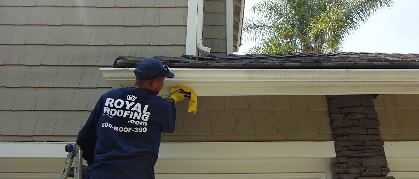 A worker in a "Royal Roofing" shirt stands on a ladder, cleaning the white gutter of a house with a yellow cloth. As a trusted roofing contractor in Orange & Los Angeles County, CA, Royal Roofing ensures every detail is spotless.