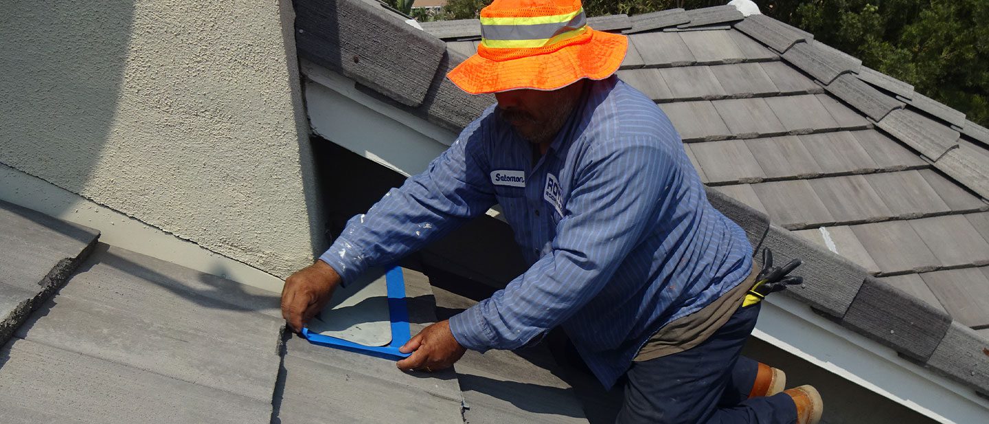 A roofing contractor in Orange & Los Angeles County, CA, kneels on a sloped roof wearing a bright orange hat and striped shirt, applying blue tape to roof tiles near a light-colored wall, surrounded by trees and neighboring rooftops.