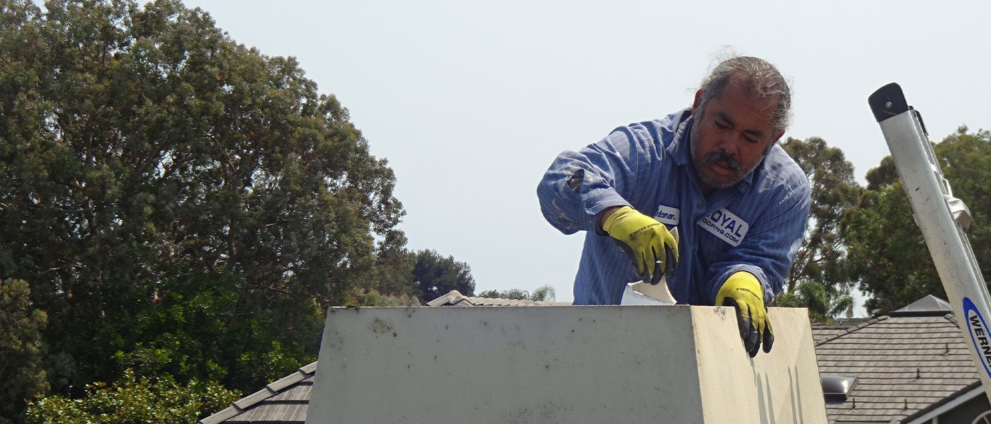 A man wearing yellow gloves and a blue shirt, likely a roofing contractor Orange & Los Angeles County, works atop a rectangular structure outdoors, performing maintenance or inspection with trees and rooftops visible in the background.