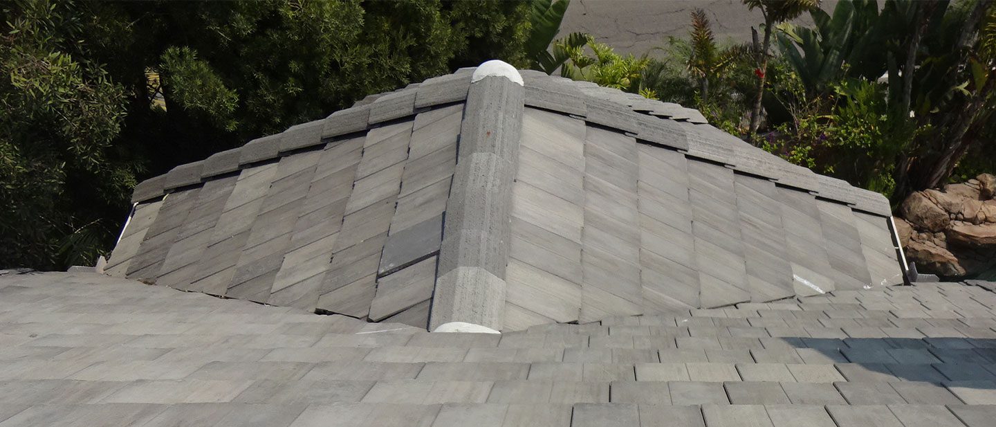 View from the peak of a gray shingle roof—expertly installed by a roofing contractor in Orange & Los Angeles County, CA—showing intersecting ridges, neat shingles, and vibrant trees and plants in the background.