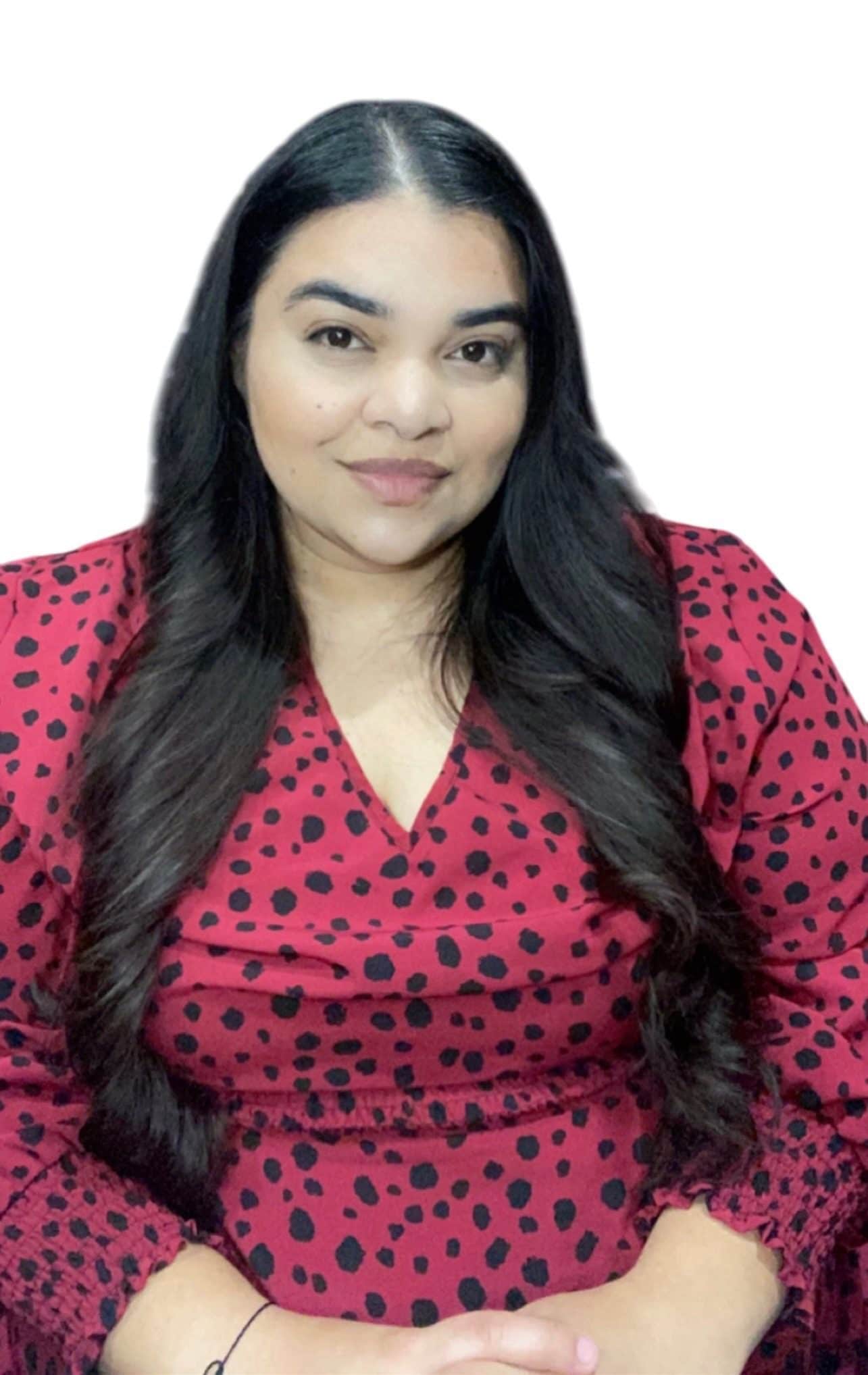 A woman with long dark hair wearing a red dress with black polka dots sits with her hands folded, smiling softly against a plain white background, much like a satisfied client of a trusted roofing contractor in Orange & Los Angeles County, CA.