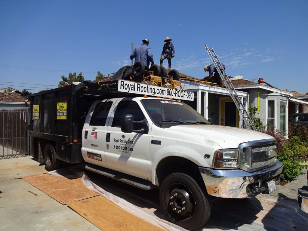 White Royal Roofing, a trusted roofing contractor in Orange & Los Angeles County, parks its truck in front of a house as workers remove shingles. Ladders, safety cones, and tools surround the scene on this bright, sunny CA day.