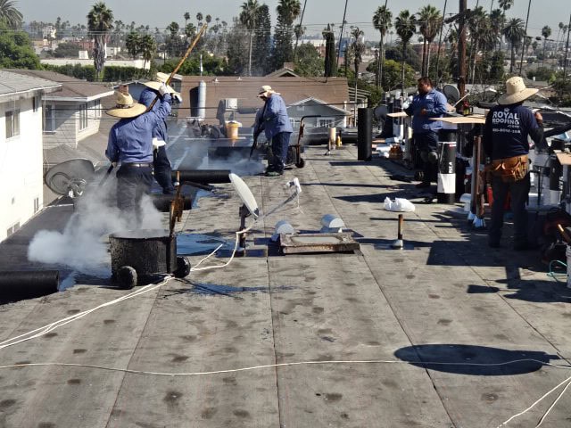 Workers wearing hats and blue shirts repair a flat rooftop in CA, using tools and materials that produce smoke. Palm trees and houses are visible in the background under a clear sky—a typical scene for a roofing contractor Orange & Los Angeles County.