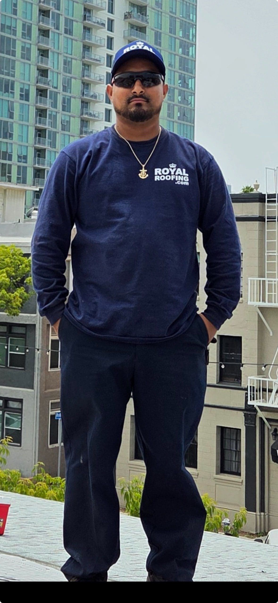 A man wearing a navy blue cap, sunglasses, a navy "Royal Roofing" sweatshirt, dark pants, and a gold anchor necklace stands outdoors in CA, representing a roofing contractor in Orange & Los Angeles County with buildings and trees behind him.