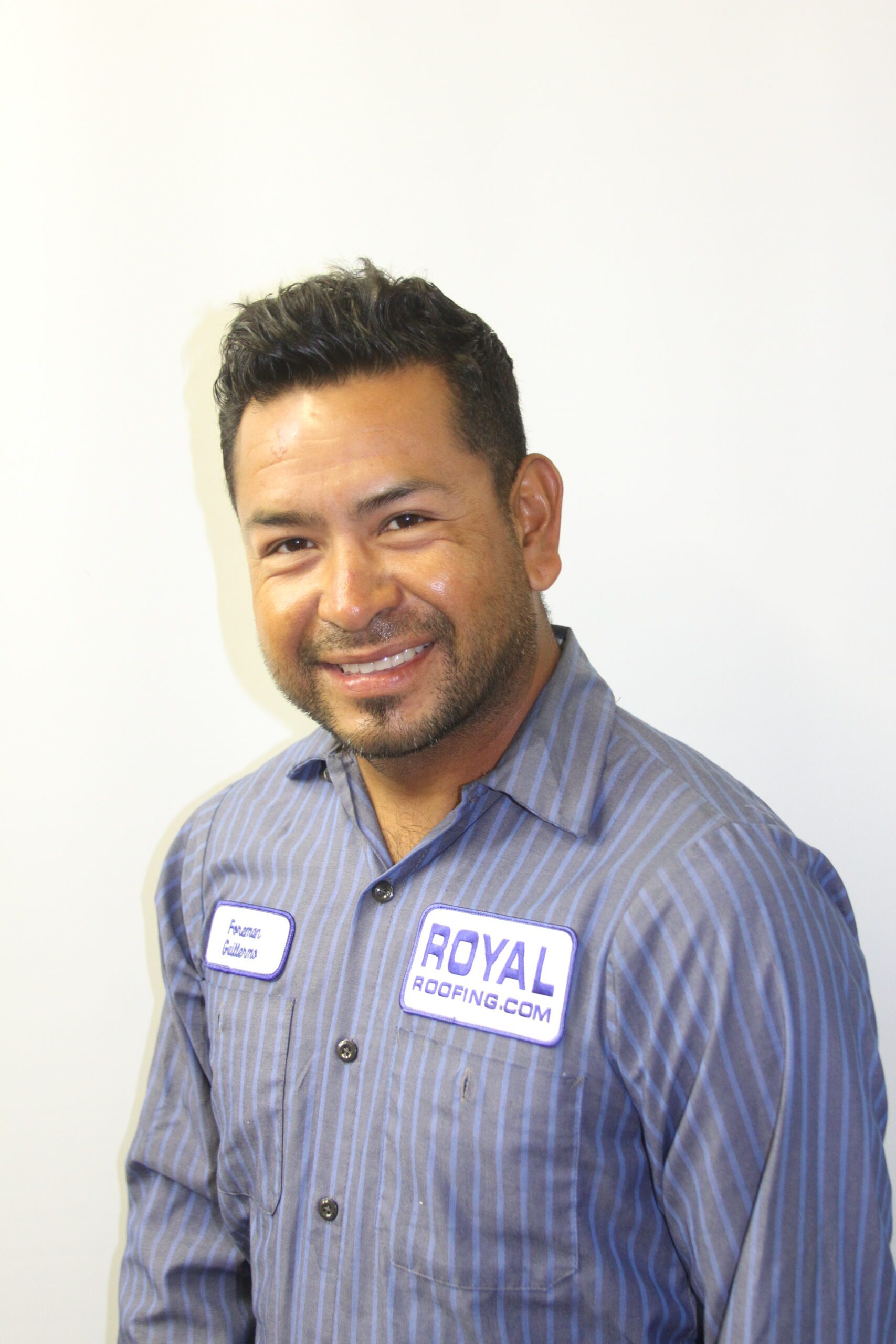 A man with short dark hair and a beard smiles at the camera, wearing a blue striped shirt with a "Royal Roofing.com" patch—a trusted roofing contractor in Orange & Los Angeles County, CA. His name tag reads "Austin." The background is plain white.
