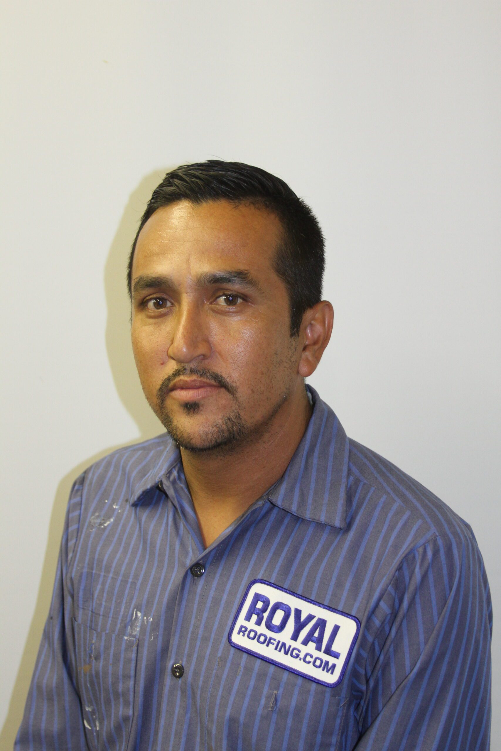 A man with short dark hair and a trimmed beard wears a blue striped shirt with a "ROYAL ROOFING.COM" patch, representing a CA roofing contractor serving Orange & Los Angeles County, sitting against a plain white background.