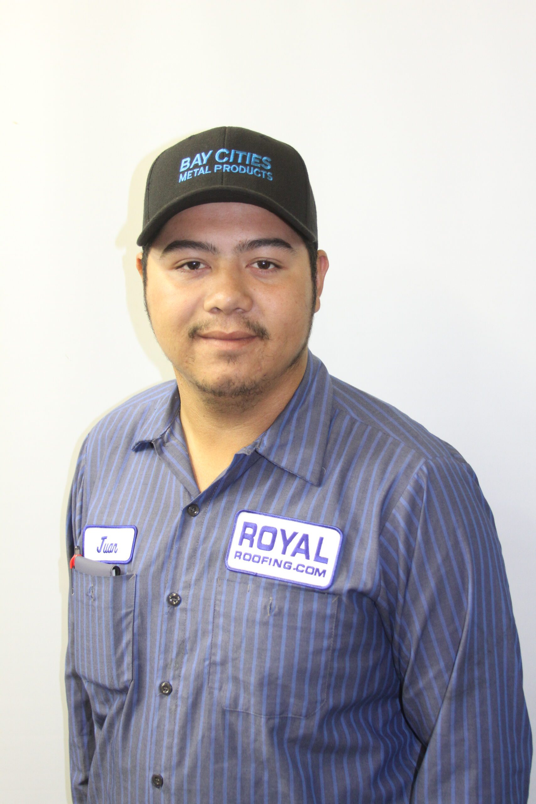 A man wearing a dark cap and a striped work shirt with patches reading “Juan” and “Royal Roofing.com” stands against a plain white background, representing a trusted roofing contractor in Orange & Los Angeles County, CA, looking at the camera with a slight smile.