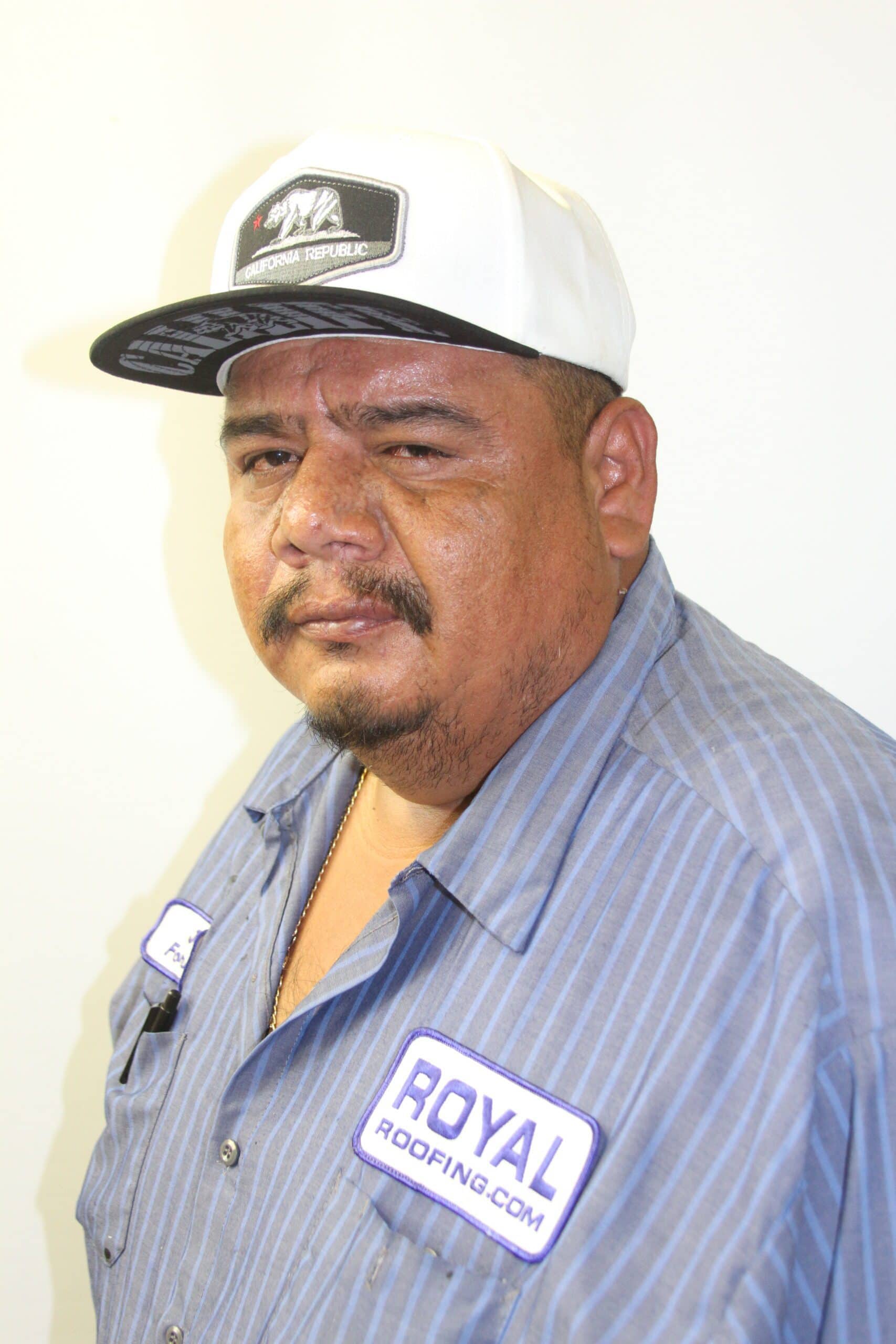 A man wearing a white and black cap, a blue button-up shirt with "Royal Roofing.com" patches, stands against a plain white background, representing a trusted roofing contractor in Orange & Los Angeles County, CA.