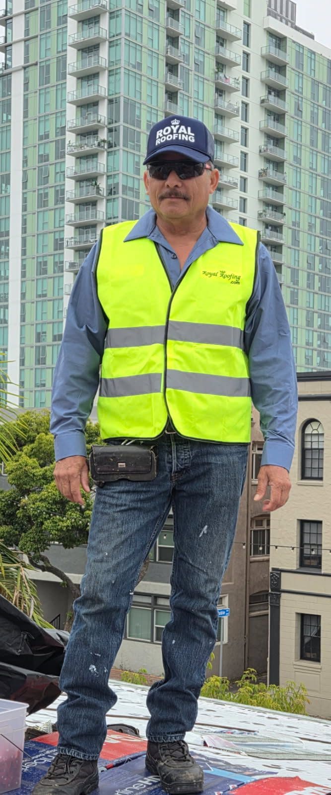 A man wearing a blue shirt, yellow safety vest, jeans, and a "Royal Moving" cap stands outdoors in CA, in front of tall buildings and trees—he looks ready for work as a roofing contractor Orange & Los Angeles County can trust. He has sunglasses and a mustache.