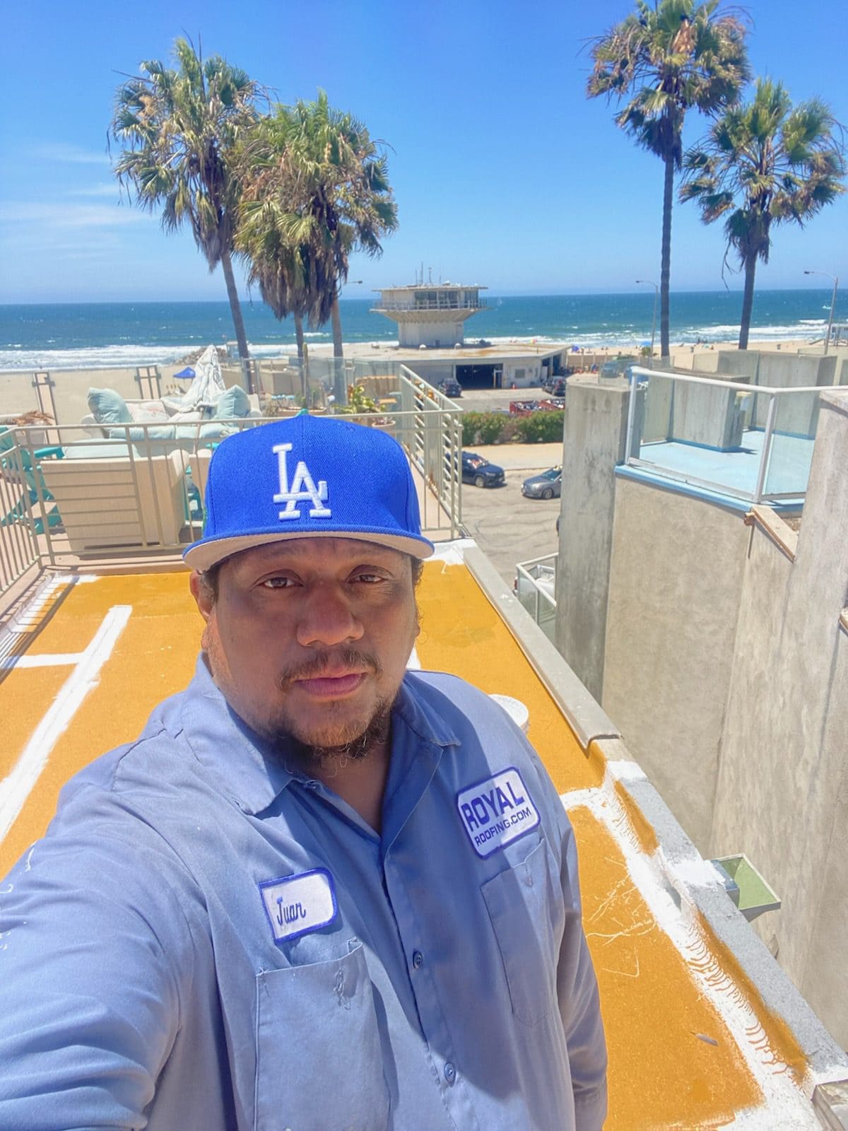 A man in a blue Los Angeles Dodgers cap and work shirt stands on a rooftop near the beach—palm trees, ocean, and a pier in the background—showcasing the expertise of a roofing contractor in Orange & Los Angeles County, CA under a sunny sky.