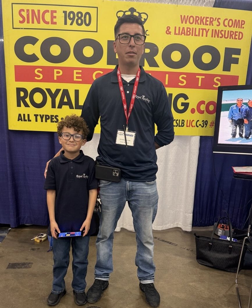 A man and a young boy in matching dark blue polo shirts and jeans smile in front of a yellow “Cool Roof Specialists” booth at an event, representing a leading roofing contractor in Orange & Los Angeles County, CA. A TV screen and banner are visible behind them.