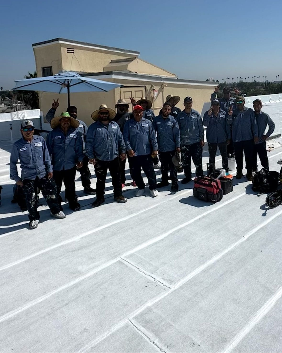 A group of workers from a roofing contractor Orange & Los Angeles County stand together on a white rooftop under a clear CA sky, wearing matching blue shirts and hats, with tools and bags nearby and a building in the background.