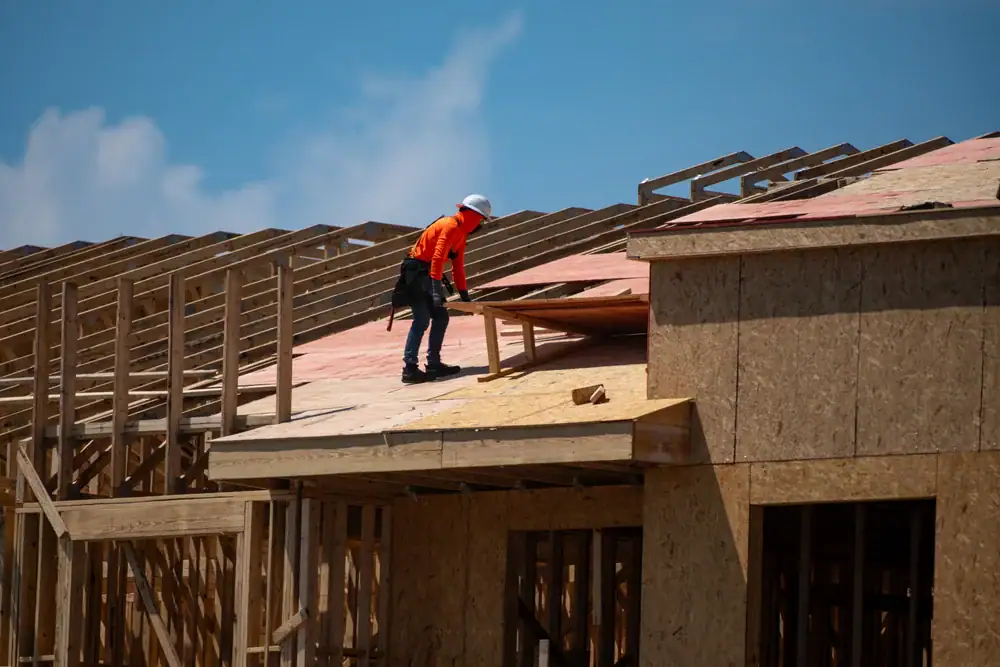 A construction worker in a safety helmet and orange shirt stands on the wooden roof frame of a building under construction, representing a skilled roofing contractor in Orange & Los Angeles County, CA, with blue sky and clouds in the background.