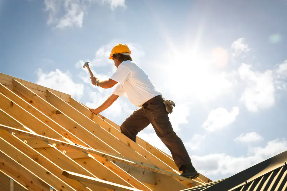 A roofing contractor in Orange & Los Angeles County, CA, wearing a yellow hard hat and white shirt, uses a hammer while building a wooden roof frame under a bright, sunny sky with scattered clouds.
