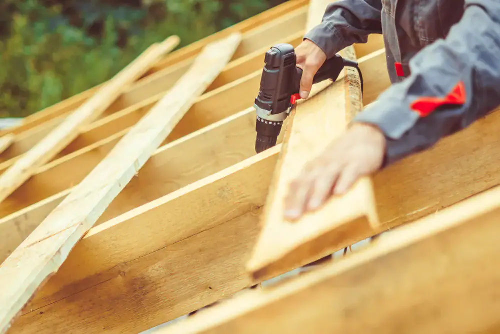 A CA roofing contractor in work clothes uses a power drill to attach wooden planks to a roof frame during construction, with greenery visible in the background—serving Orange & Los Angeles County.