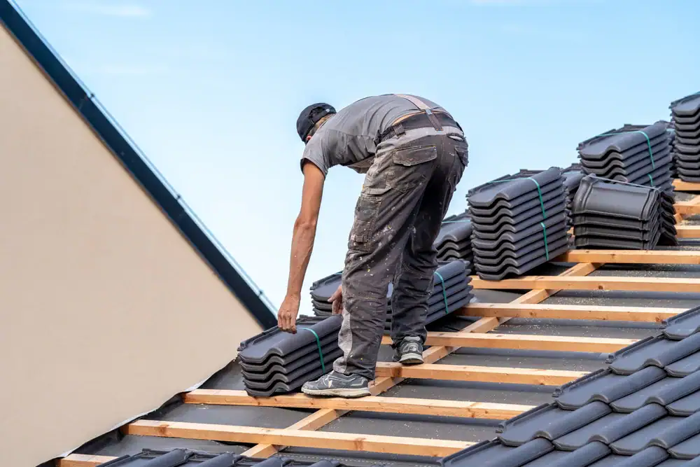 A construction worker in overalls, employed by a roofing contractor Orange & Los Angeles County, installs roof tiles on a wooden frame. Stacks of black tiles are lined up, and the sky is clear and blue in the background.