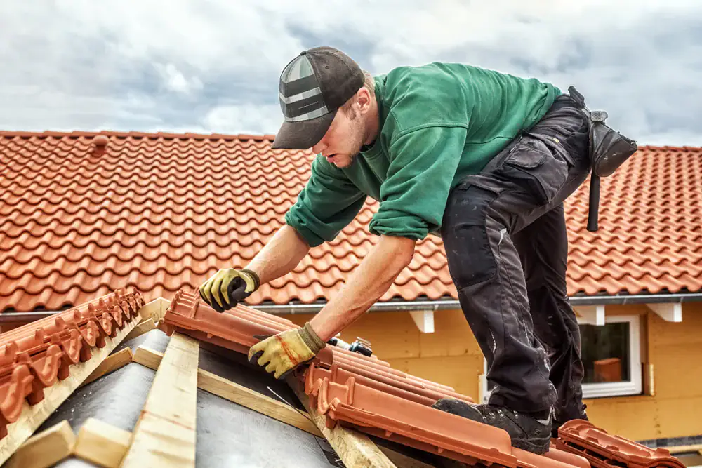 A roofing contractor Orange & Los Angeles County, wearing a green sweatshirt, black pants, gloves, and a cap, installs red roof tiles on a house’s sloped rooftop under a cloudy CA sky.