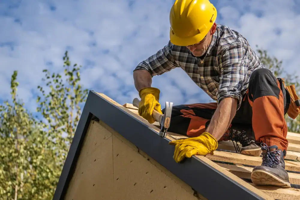 A construction worker in a yellow hard hat and gloves, representing a roofing contractor Orange & Los Angeles County, is nailing a board on a sloped wooden roof under a blue sky, surrounded by trees.