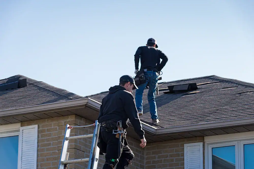 Two workers wearing safety gear stand on the roof of a house near a ladder, as a roofing contractor Orange & Los Angeles County team inspects or repairs the shingles under a clear blue CA sky.