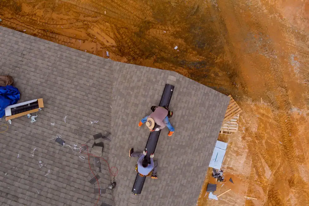 Aerial view of two workers from a roofing contractor in Orange & Los Angeles County, CA, installing a vent on the peaked roof of a house under construction, with tools and building materials scattered on the ground below.