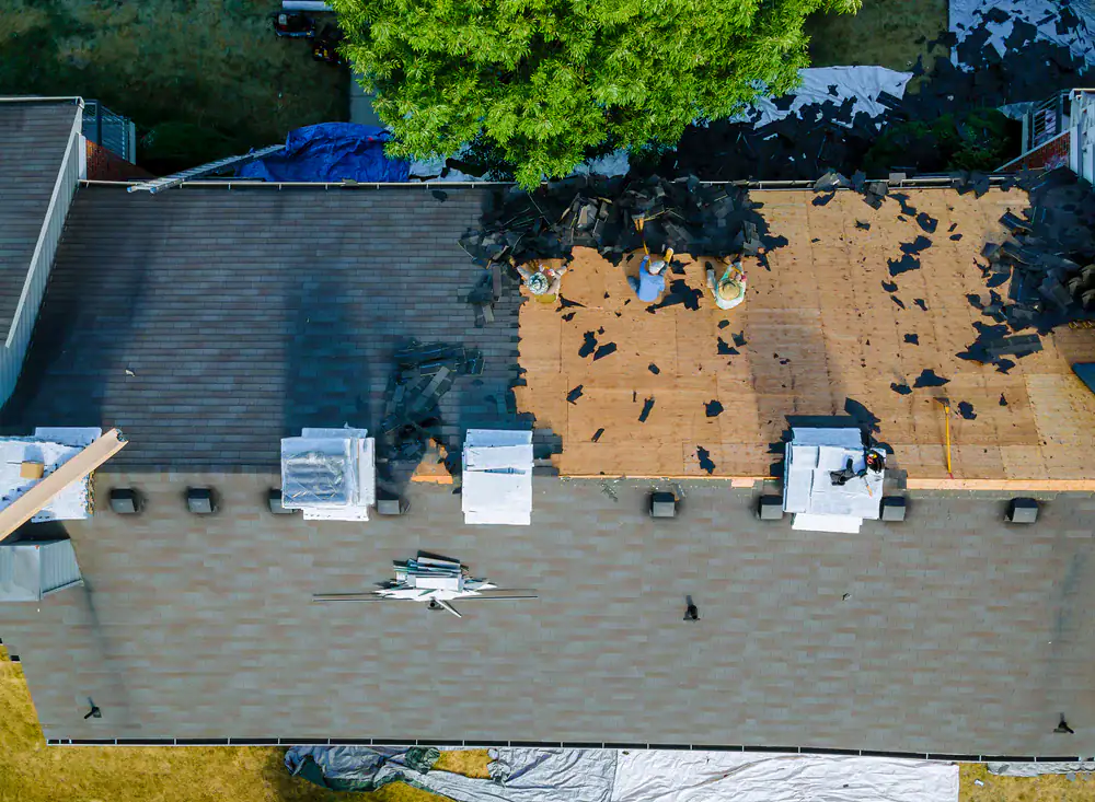 Aerial view of workers from a CA roofing contractor Orange & Los Angeles County removing old shingles, with debris scattered and a tree partially covering the area. The roof is partially stripped, exposing wooden panels beneath.