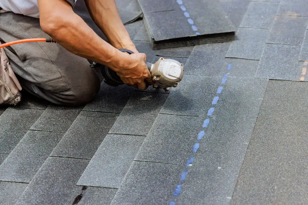 A roofing contractor Orange & Los Angeles County uses a power tool to install or repair dark asphalt shingles, kneeling on the rooftop with several overlapping shingles visible.