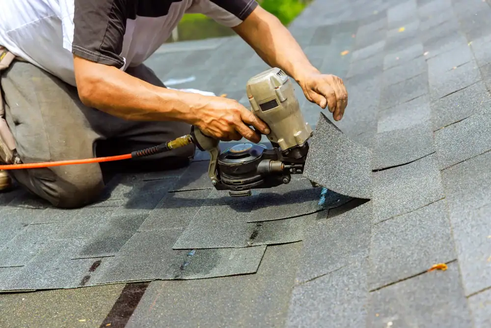 A roofing contractor in Orange & Los Angeles County, CA, installs asphalt shingles on a roof using a pneumatic nail gun, carefully kneeling and aligning each shingle before securing it in place.