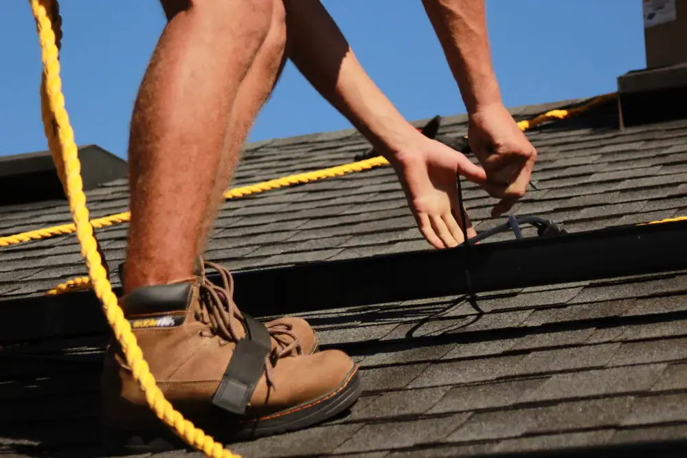 A roofing contractor in Orange & Los Angeles County, CA, wearing brown work boots, installs equipment on a shingled roof and secures black wires. Yellow safety ropes are visible, with a clear blue sky in the background.