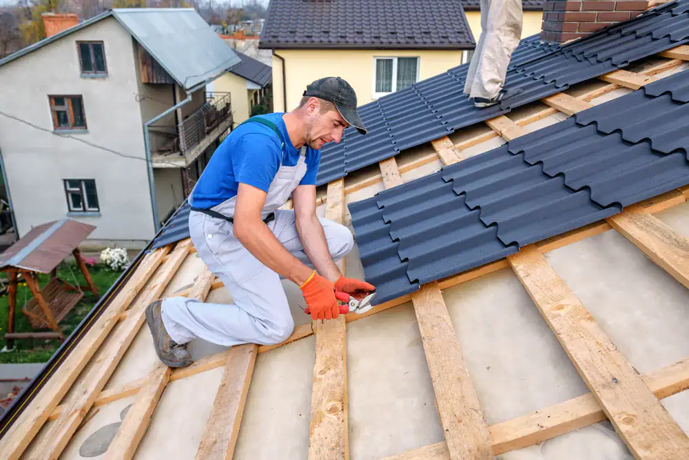 A construction worker in overalls and gloves, likely a roofing contractor in Orange & Los Angeles County, CA, installs metal roofing sheets on a house roof with pliers. Another person stands nearby. Houses and a swing set are visible in the background.