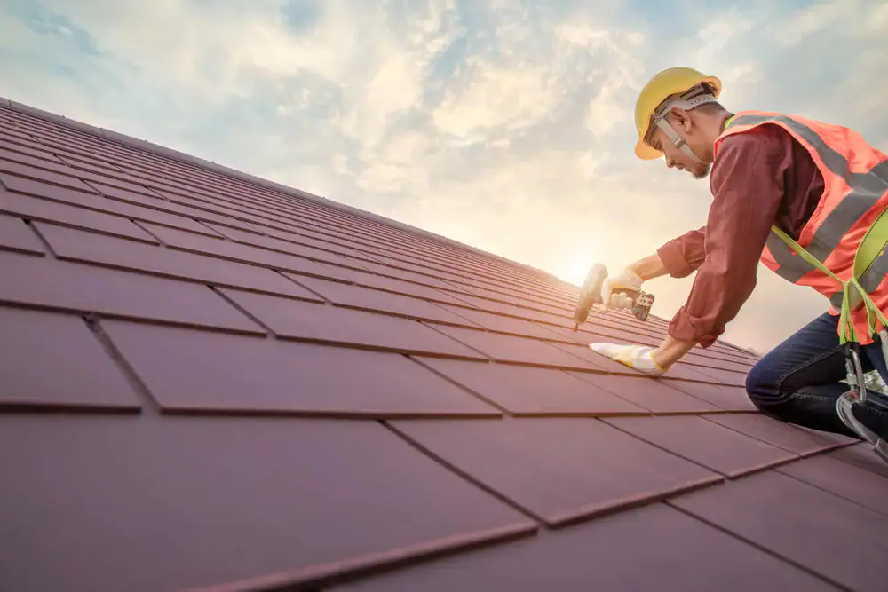 A roofing contractor in Orange & Los Angeles County, CA, wearing a yellow helmet and safety vest, kneels on a sloped roof, installing tiles with a power drill as the sun sets in the background.