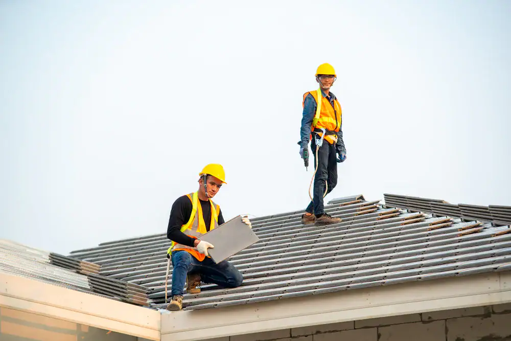 Two construction workers wearing yellow safety helmets and orange vests are installing tiles on a roof. One kneels, holding a tile, while the other stands and smiles. A trusted roofing contractor Orange & Los Angeles County, CA, ensures quality work.