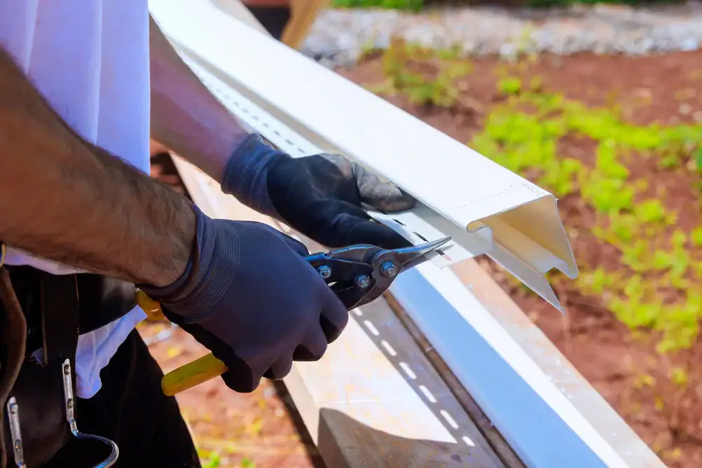 Worker cutting white metal rain gutter with hand shears during gutter installation on a residential building exterior.