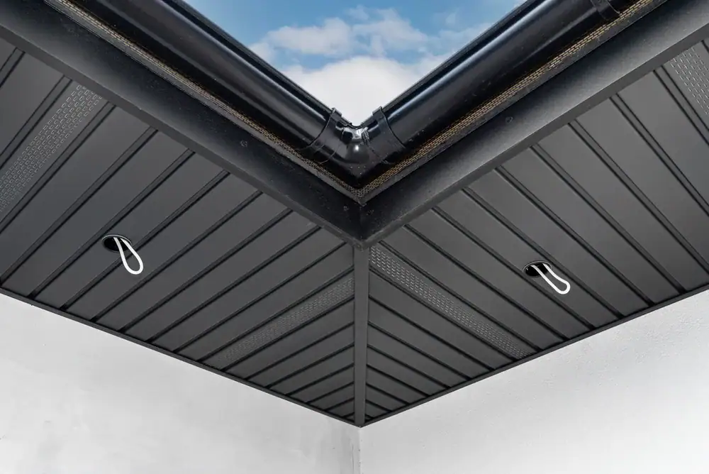 Modern house roof soffit corner with black rain gutters, recessed ceiling lights, and dark metal cladding under a blue sky.
