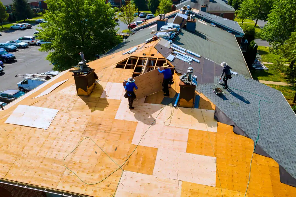 Aerial view of workers repairing a roof, placing new plywood sheets on a large building. Construction tools and materials are spread around, and trees and parked cars are visible in the background.
