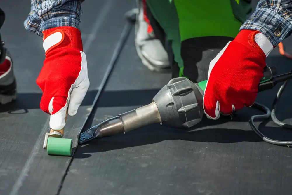 Close-up of a worker wearing red gloves using a heat gun and a green roller to seal seams on a flat black roofing surface.